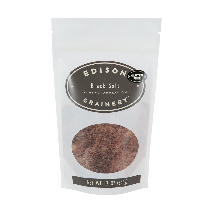 A 12 oz bag of Black Salt standing upright in a bio-degradable bag. A grey oval label, bearing the product name sits above an oval viewing window revealing the product.