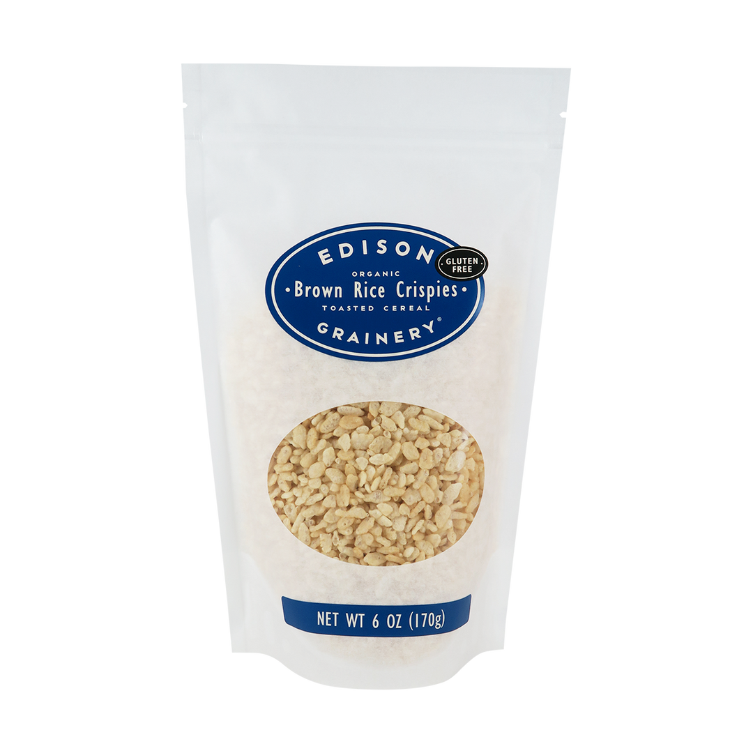 A 6 oz bag of Organic Brown Rice Crispies standing upright in a bio-degradable bag. A royal blue, oval label bearing the product name sits above an oval viewing window revealing the product.