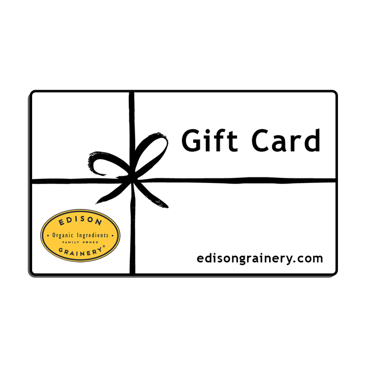 A white rectangular gift wrapped in a black ribbon with the words Gift Card, edisongrainery.com and a golden yellow logo superimposed atop it.
