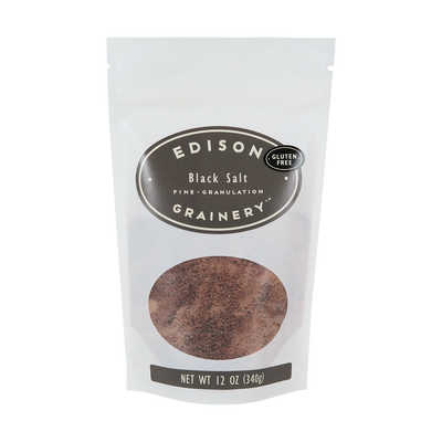 A 12 oz bag of Black Salt standing upright in a bio-degradable bag. A grey oval label, bearing the product name sits above an oval viewing window revealing the product.