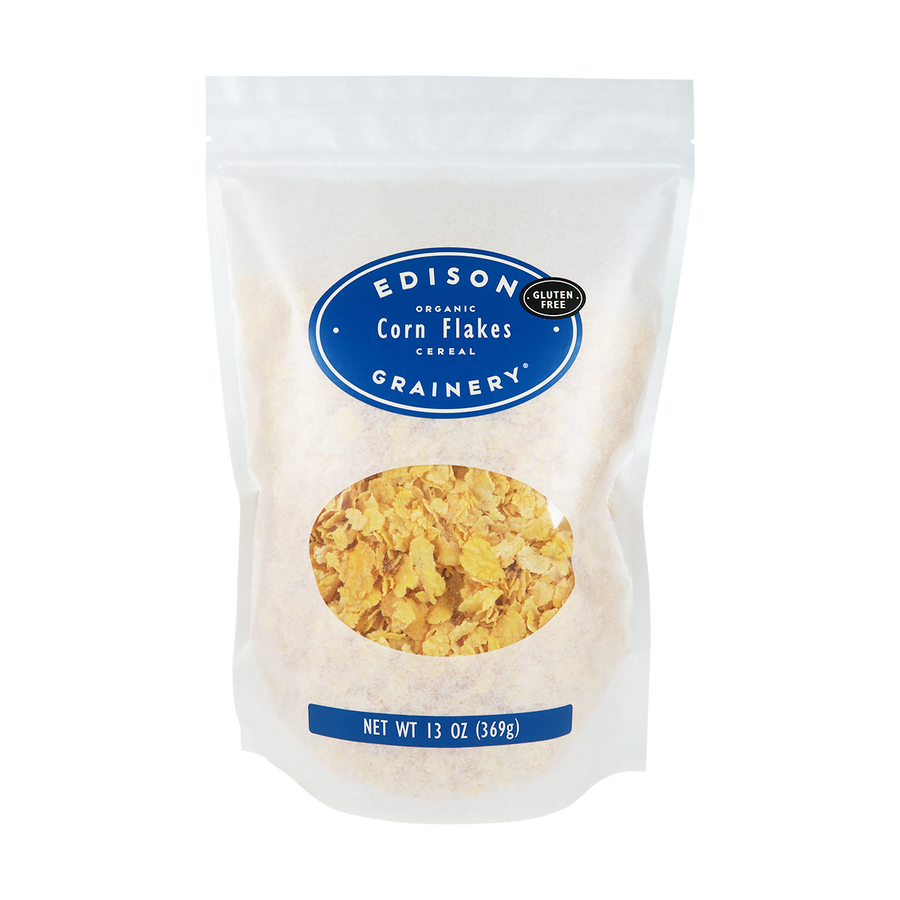 A 13 oz bag of Organic Corn Flakes Cereal standing upright in a bio-degradable bag. A royal blue, oval label bearing the product name sits above an oval viewing window revealing the product.