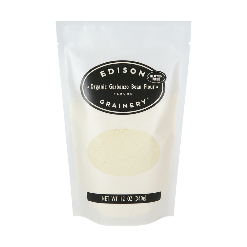 A 12 oz bag of Organic Garbanzo Bean Flour standing upright in a bio-degradable bag. A black oval label, bearing the product name sits above an oval viewing window revealing the product.