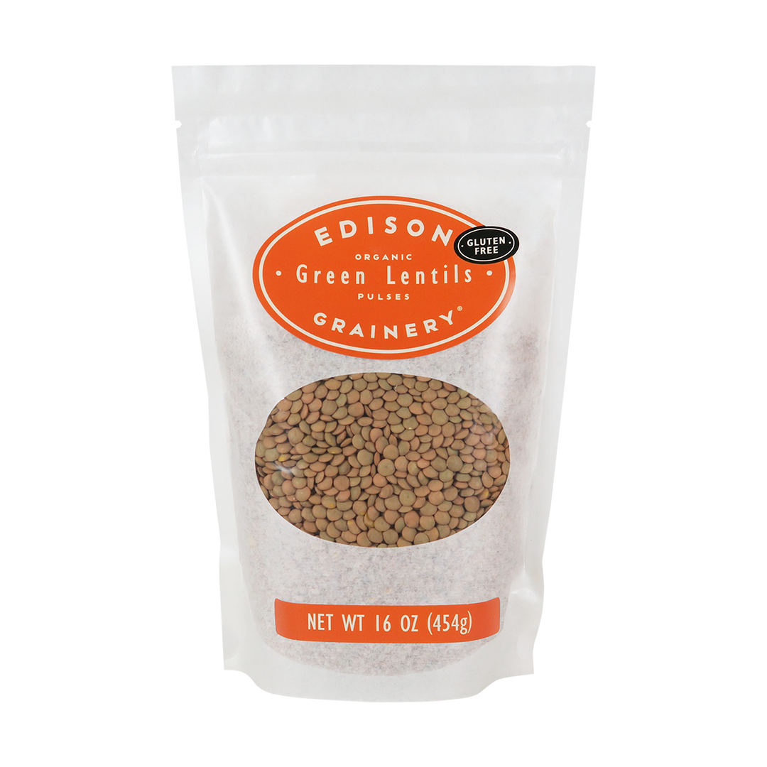 A 16 oz bag of Organic Green Lentils standing upright in a bio-degradable bag. An orange oval label, bearing the product name sits above an oval viewing window revealing the product.