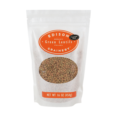 A 16 oz bag of Organic Green Lentils standing upright in a bio-degradable bag. An orange oval label, bearing the product name sits above an oval viewing window revealing the product.