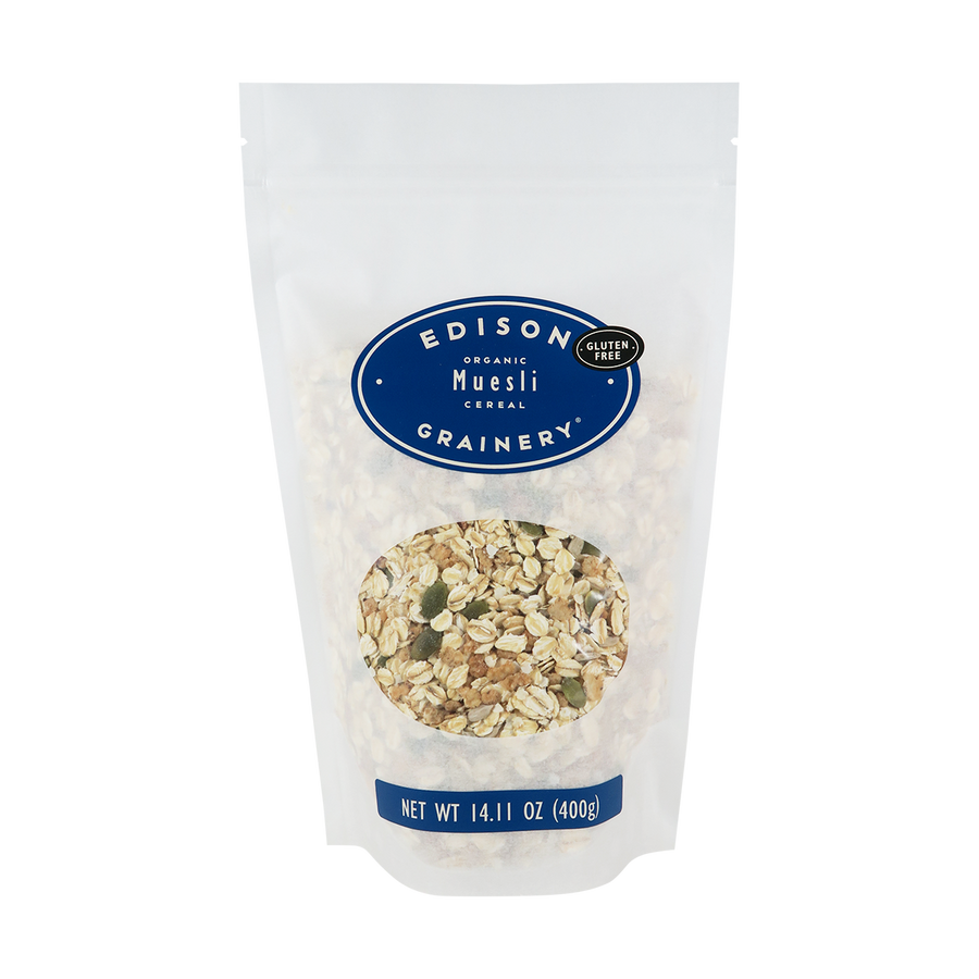 A 14.11 oz bag of Organic Muesli standing upright in a bio-degradable bag. A royal blue oval label, bearing the product name sits above an oval viewing window revealing the product.