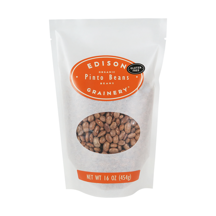 A 16 oz bag of Organic Pinto Beans standing upright in a bio-degradable bag. An orange oval label, bearing the product name sits above an oval viewing window revealing the product.