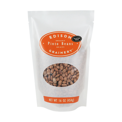A 16 oz bag of Organic Pinto Beans standing upright in a bio-degradable bag. An orange oval label, bearing the product name sits above an oval viewing window revealing the product.