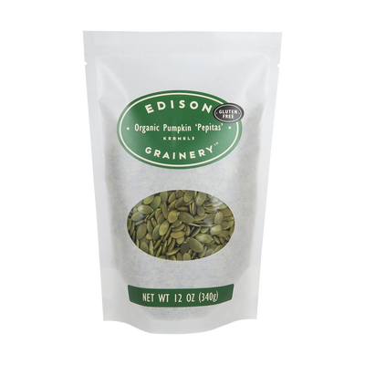 A 12 oz bag of Organic Pumpkin 'Pepitas" standing upright in a bio-degradable bag. A forest green oval label, bearing the product name sits above an oval viewing window revealing the product.