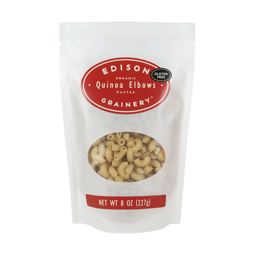 An 8 oz bag of Organic Quinoa Pasta: Elbows standing upright in a bio-degradable bag. A crimson red oval label, bearing the product name sits above an oval viewing window revealing the product.