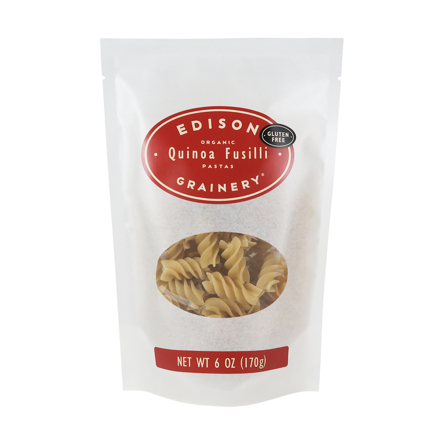A 6 oz bag of Organic Quinoa Pasta: Fusilli standing upright in a bio-degradable bag. A crimson red oval label, bearing the product name sits above an oval viewing window revealing the product.