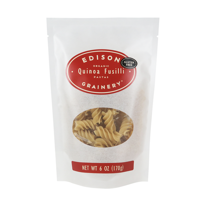 A 6 oz bag of Organic Quinoa Pasta: Fusilli standing upright in a bio-degradable bag. A crimson red oval label, bearing the product name sits above an oval viewing window revealing the product.