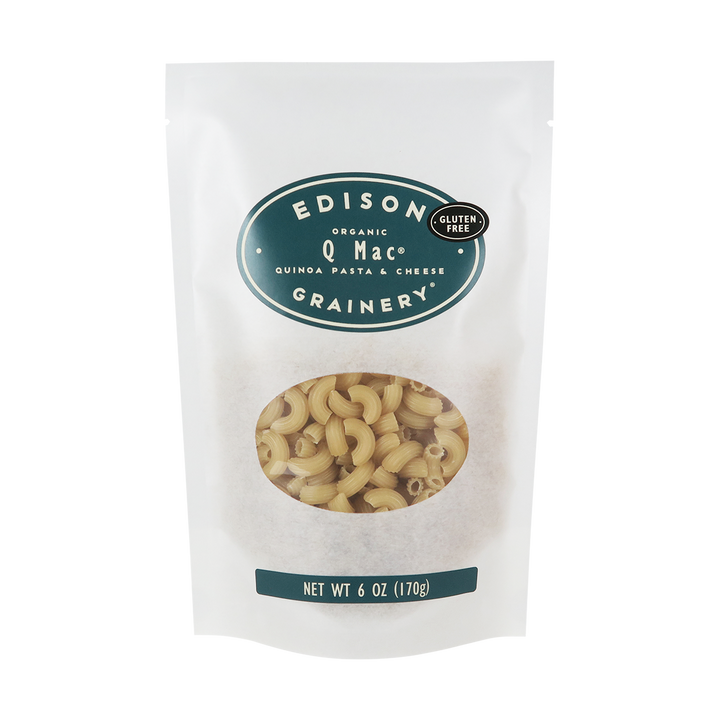 A 6 oz bag of Organic Q-Mac standing upright in a bio-degradable bag. A dark teal oval label, bearing the product name sits above an oval viewing window revealing the product.