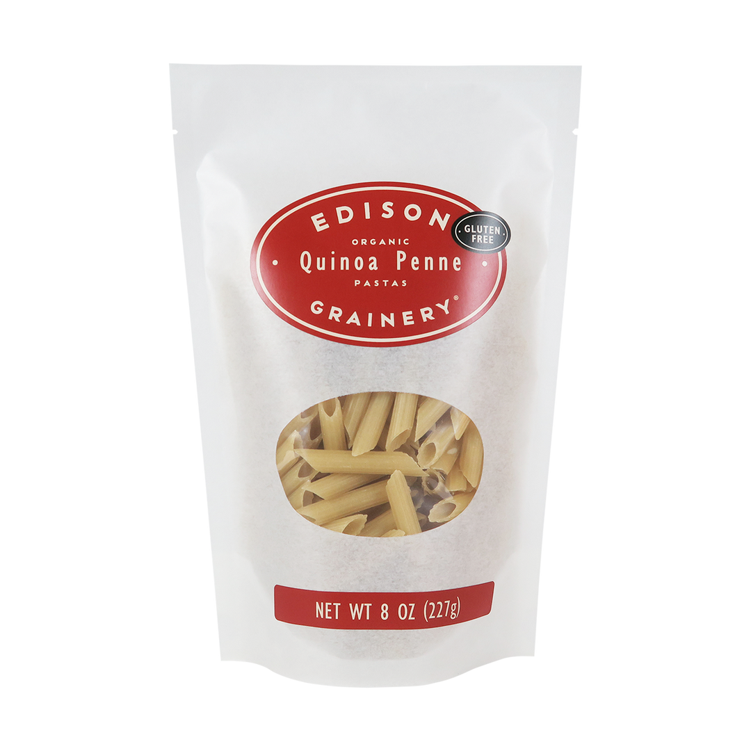 An 8 oz bag of Organic Quinoa Pasta: Penne standing upright in a bio-degradable bag. A crimson red oval label, bearing the product name sits above an oval viewing window revealing the product.
