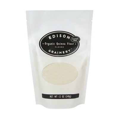 A 12 oz bag of Organic Quinoa Flour standing upright in a bio-degradable bag. A black oval label, bearing the product name sits above an oval viewing window revealing the product.