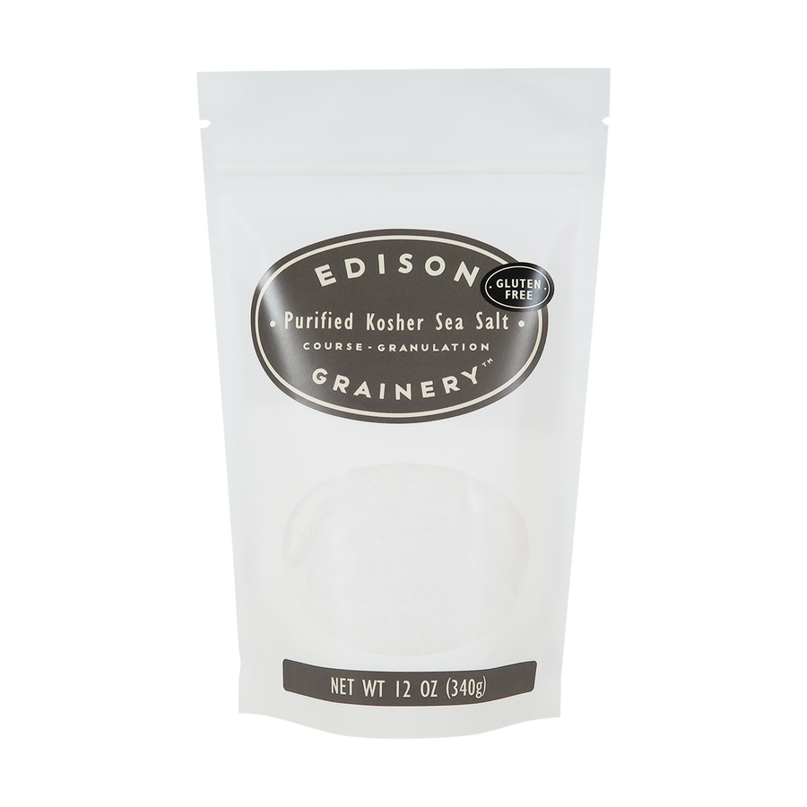 A 12 oz bag of Kosher Sea Salt standing upright in a bio-degradable bag. A grey oval label, bearing the product name sits above an oval viewing window revealing the product.