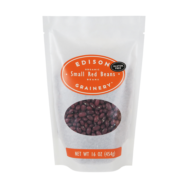 A 16 oz bag of Organic Small Red Beans standing upright in a bio-degradable bag. An orange oval label, bearing the product name sits above an oval viewing window revealing the product.