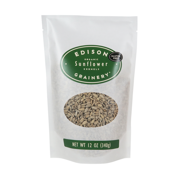 A 12 oz bag of Organic Sunflower Kernels standing upright in a bio-degradable bag. A forest green oval label, bearing the product name sits above an oval viewing window revealing the product.