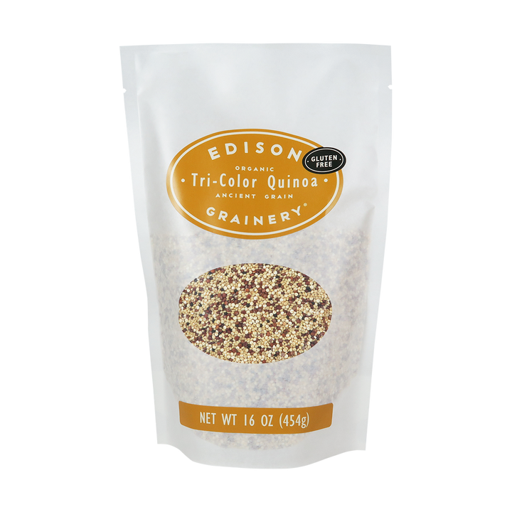 A 16 oz bag of Organic Tri-Color Quinoa standing upright in a bio-degradable bag. A golden yellow oval label, bearing the product name sits above an oval viewing window revealing the product.