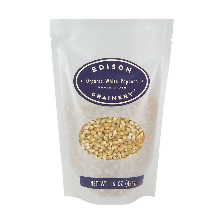 A 16 oz bag of Organic White Popcorn standing upright in a bio-degradable bag. A deep purple oval label, bearing the product name sits above an oval viewing window revealing the product.