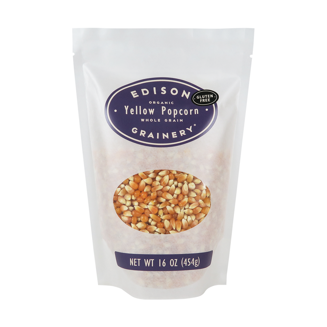 A 16 oz bag of Organic Yellow Popcorn standing upright in a bio-degradable bag. A deep purple oval label, bearing the product name sits above an oval viewing window revealing the product.