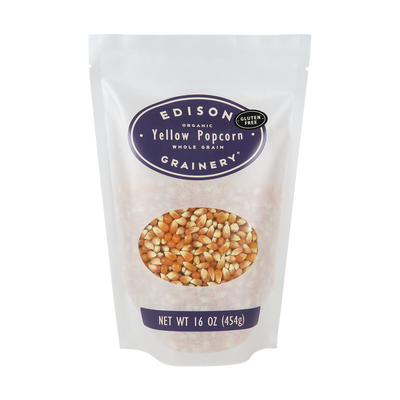 A 16 oz bag of Organic Yellow Popcorn standing upright in a bio-degradable bag. A deep purple oval label, bearing the product name sits above an oval viewing window revealing the product.